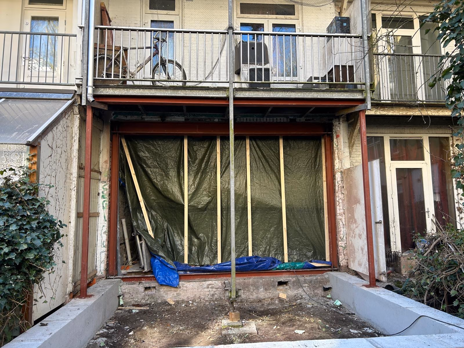 Achtergevel met stalen portaal, houten regelwerk en afdekzeil in de nieuwe opening onder een balkon